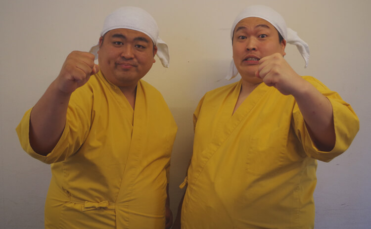 黄色い衣装と白い頭巾姿で、カメラに向けて拳を出すゆんぼだんぷのふたり。